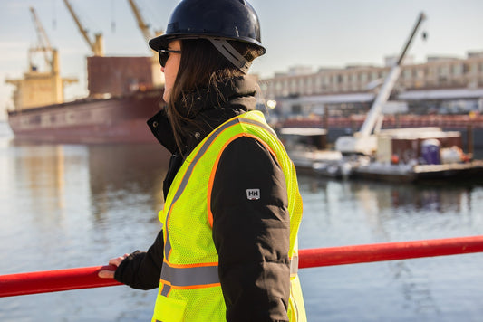 Female worker wearing an ARX high-visibility safety vest and hard hat overlooking an industrial waterfront worksite.