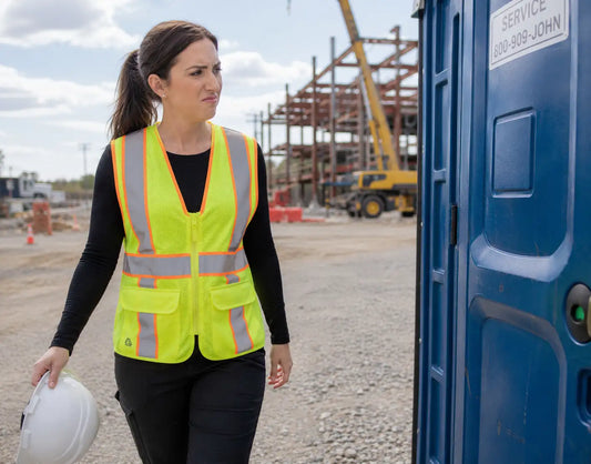 Woman construction worker in an ARX safety vest walking toward a blue porta-potty holding a hard hat on a construction site